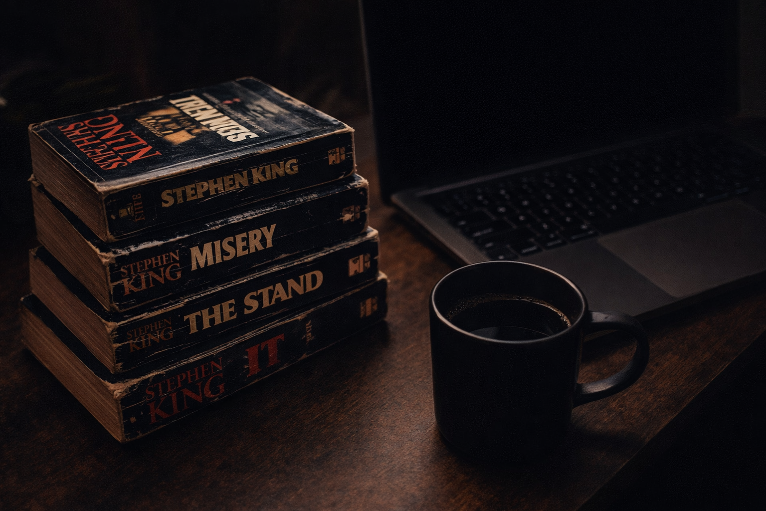 A stack of Stephen King horror novels on an industrial wooden desk with a laptop and coffee cup